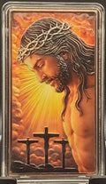 A small rectangular religious plaque or card in a clear plastic case, shown on a black display stand. The artwork depicts Jesus with a crown of thorns, radiant golden light, clouds, and three crosses against a dark studio background.