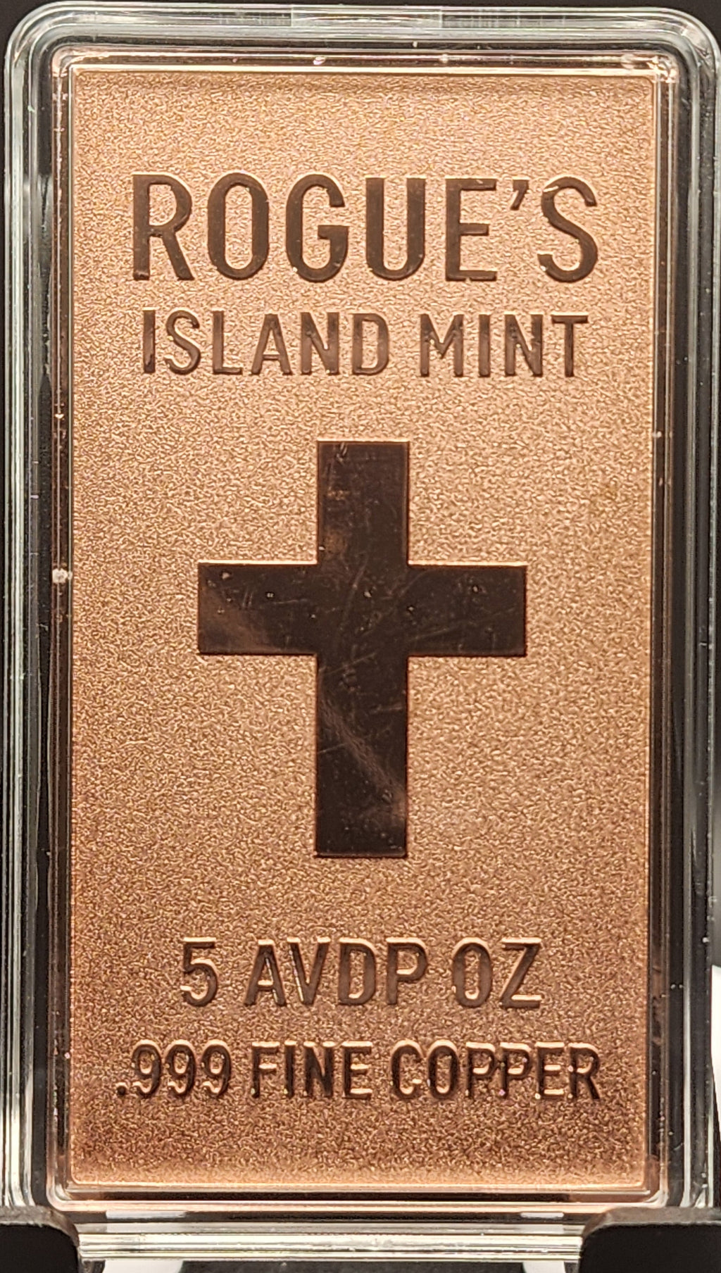 A rectangular copper-colored metal bar in a clear plastic case, labeled 'ROGUE'S ISLAND MINT' with a large cross emblem and text reading '5 AVDP OZ .999 FINE COPPER,' displayed on a black stand against a dark background.