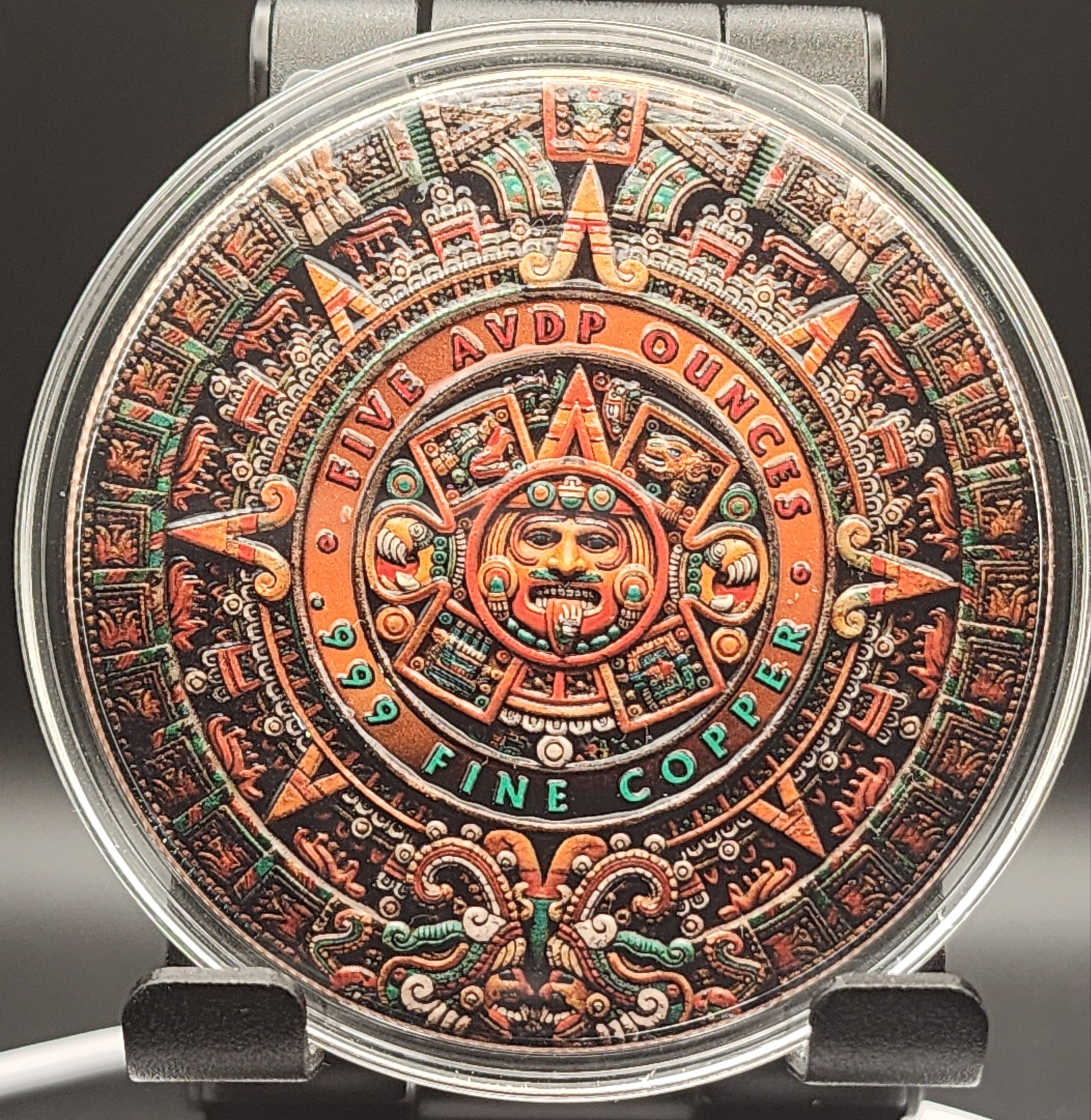 A close-up of a round watch dial featuring an intricate Aztec calendar design in copper, orange, green, and black, with a silver-toned metal case and dark strap.
