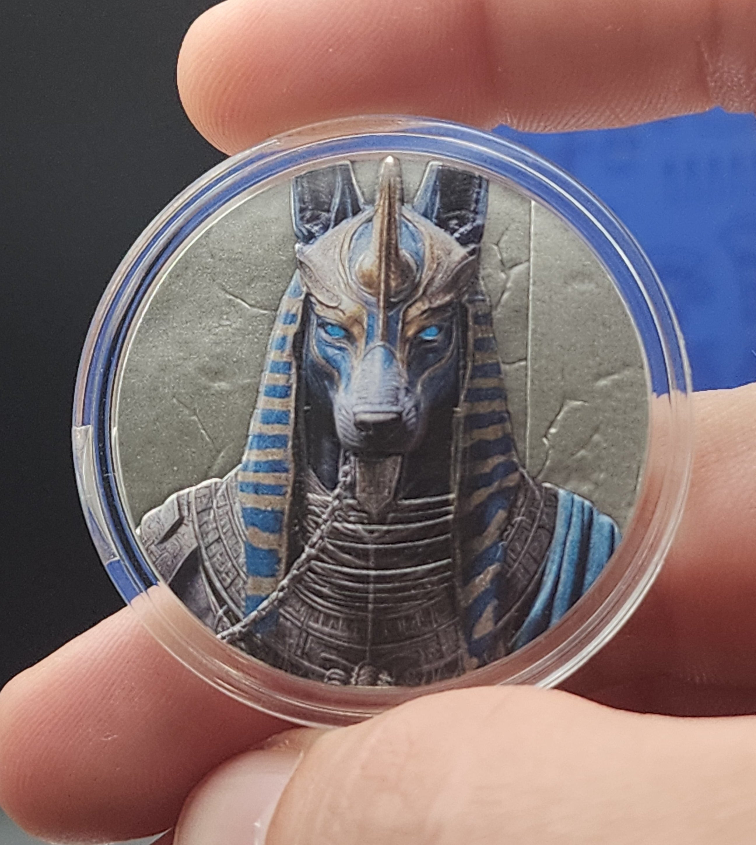 A close-up of a hand holding a round clear plastic capsule containing an illustrated figure with blue, gray, and black details against a dark background.