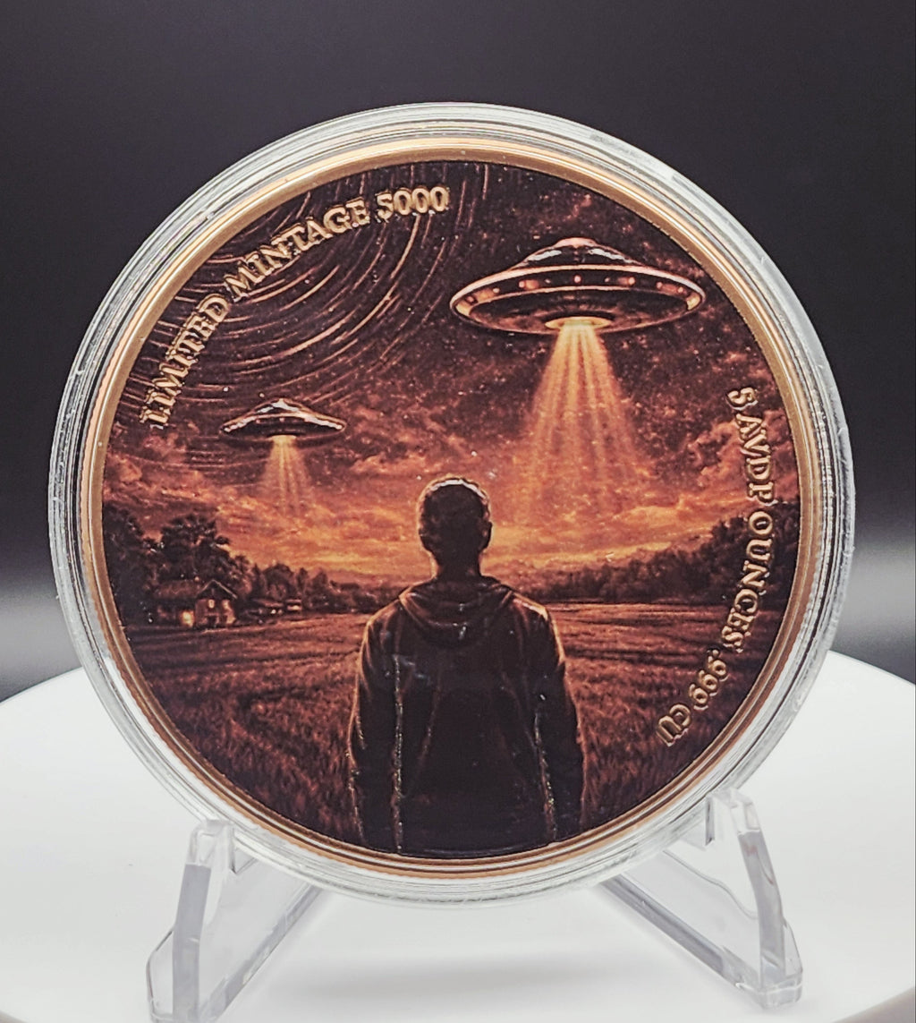 A round commemorative coin in a clear protective case, displayed on a clear stand against a dark background. The coin features an illustration of a person standing in a field with two UFOs emitting beams of light in an orange and brown sunset sky.