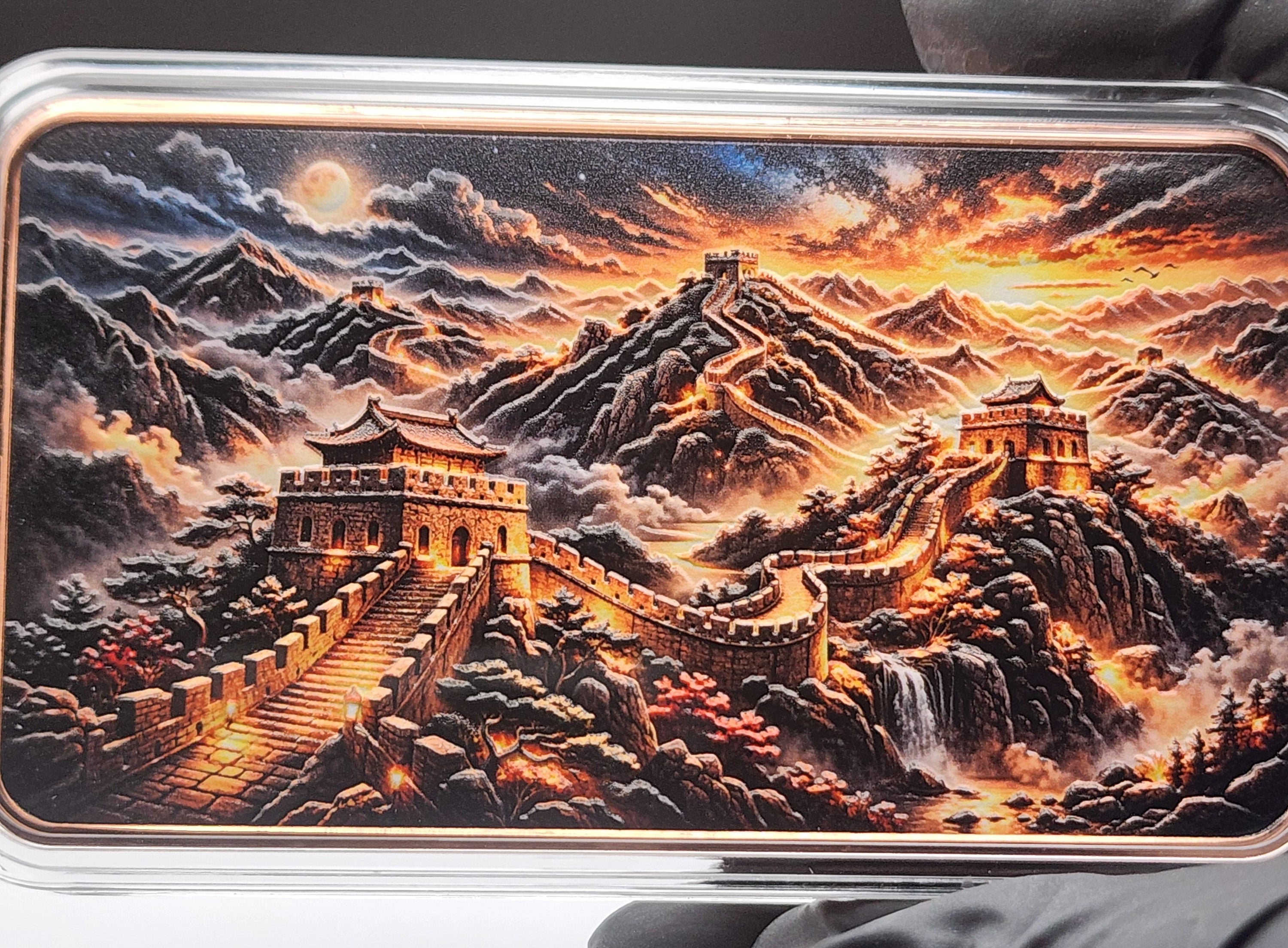 A rectangular metal bar or coin with a detailed landscape print of the Great Wall of China, held by black-gloved hands over a white circular platform against a dark background.